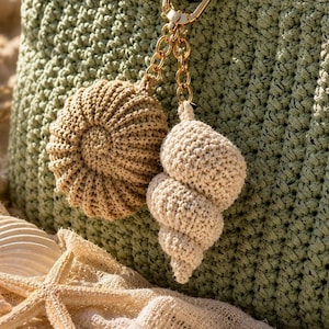 Puede incluir: Un bolso de ganchillo color verde salvia con un llavero de metal dorado que presenta dos colgantes tejidos en forma de concha espiral y caracola, en tonos beige y bronce, sujetos al asa trenzada del bolso.