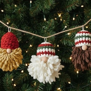 May include: Three handmade Christmas gnome ornaments hanging on a string. Each gnome has a crocheted hat in red, white, and green, with a matching beard. The beards are in shades of yellow, white, and brown. The ornaments are set against a backdrop of a Christmas tree.