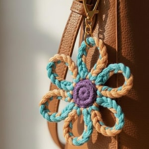 May include: A crochet flower keyring in shades of blue, tan, and purple. The flower is attached to a gold-coloured clasp and ring, hanging against a brown leather bag. The flower's petals are a mix of blue and tan yarn, creating a decorative accessory.