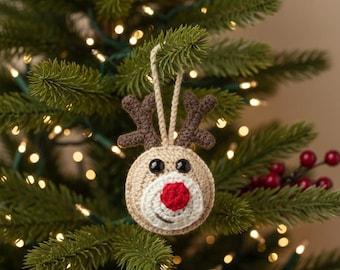 May include: A handmade Christmas tree ornament shaped like a reindeer. The ornament is crocheted with tan yarn, featuring brown antlers, black eyes, and a red nose. It hangs from a loop of matching yarn, displayed on a Christmas tree.