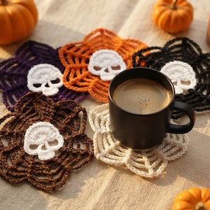 May include: Five crocheted coasters in purple, orange, black, brown, and cream, each featuring a central white skull motif and a web-like border. A black ceramic mug sits on the cream coaster, surrounded by small orange pumpkins and a candle.