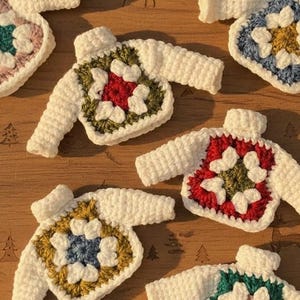 Puede incluir: Una colección de adornos de suéter de ganchillo hechos a mano en varios colores, incluyendo rojo, verde, azul y amarillo, con detalles blancos. Los suéteres están dispuestos sobre una superficie de madera. El texto "KnitsByNeseArt" es visible.