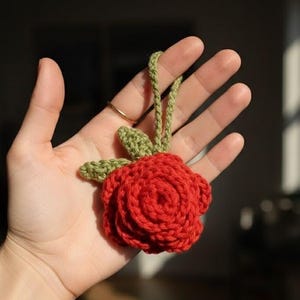 Può includere: Una rosa all'uncinetto fatta a mano in rosso vibrante, con foglie verdi e un anello verde per appenderla. Il fiore è tenuto in una mano, mostrando i dettagli della cucitura e la consistenza del filato. Un elemento floreale decorativo.