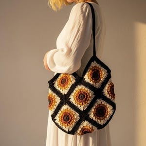 May include: A black and white crochet tote bag with a sunflower pattern. The bag features a repeating pattern of yellow, orange, and white sunflowers. The bag has a black strap and is held by a person wearing a white dress.