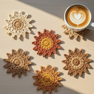 May include: Six handmade crochet sunburst coasters in shades of brown, beige, and orange. One coaster supports a white mug with latte art. The coasters are arranged on a light-colored surface.