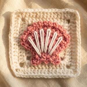 May include: A square crochet granny square featuring a central motif of a pink seashell with white embroidered vertical lines. The square is crafted from off-white yarn with a textured border, showcasing detailed stitching and a raised shell design.
