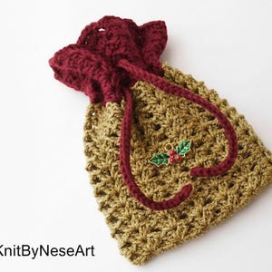 Puede incluir: Una bolsa con cordón hecha a mano de ganchillo en oro y burdeos. La bolsa presenta un patrón de puntadas texturizadas y un cierre de cordón burdeos. Un pequeño dije de acebo con bayas rojas y hojas verdes está unido al cordón. La bolsa está etiquetada como "KnitByNeseArt".