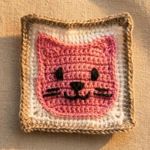 May include: A square crochet coaster featuring a pink cat face centered on a white background with a tan border. The cat face is crafted with pink yarn and includes black embroidered eyes, a small black nose, and whiskers in a simple design.