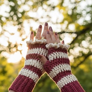 Könnte beinhalten: Gehäkelte fingerlose Handschuhe in bordeauxroten und cremefarbenen Streifen. Die Handschuhe haben eine gerüschte cremefarbene Bordüre am Handgelenk. Das Bild ist vor einem verschwommenen grün-gelben Hintergrund.