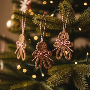 Puede incluir: Adornos navideños de hombre de jengibre de ganchillo. Los adornos marrones cuelgan de un árbol de Navidad. Cada adorno tiene un lazo a rayas rojas y blancas y una cuerda a juego para colgar. El árbol está decorado con luces.
