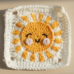 May include: A crocheted square featuring a smiling sun design. The sun is yellow with a black smiling face and pink cheeks, surrounded by yellow rays. The square is bordered in white.