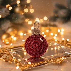 May include: A handcrafted, red crocheted Christmas ornament with a spiral design. The ornament has a tan top and a loop for hanging. It is displayed on a gold-framed mirror with blurred fairy lights in the background, creating a festive holiday scene.