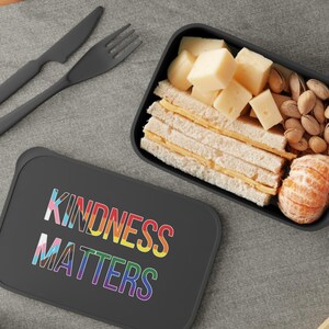 Może przedstawiać: Czarny pojemnik na lunch z napisem "KINDNESS MATTERS" w tęczowych kolorach. Pojemnik zawiera kanapki, ser, orzechy i mandarynkę. Obok leżą czarny widelec i nóż.