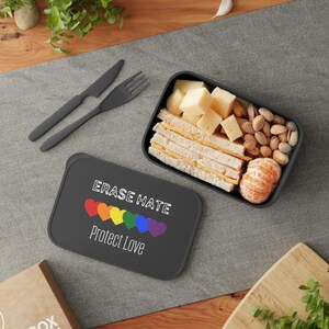 May include: A black lunchbox with the text "ERASE HATE Protect Love" and a rainbow heart design. The lunchbox is open, revealing sandwiches, cheese cubes, nuts, and a peeled mandarin orange. A fork and knife are next to the lunchbox.