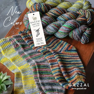 May include: A wooden surface displays colourful hand-painted yarn skeins and striped socks. A tag reads "GAZZAL HAPPY FEET Hand Painted" with yarn composition details. The socks and yarn feature a mix of colours including yellow, blue, orange, and green.