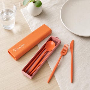 Peut inclure: Un ensemble de couverts orange portable comprenant une cuillère, une fourchette et un couteau. L'étui rectangulaire orange porte le nom Penelope imprimé en écriture blanche. La fourchette et le couteau sont posés à côté de l'étui ouvert.