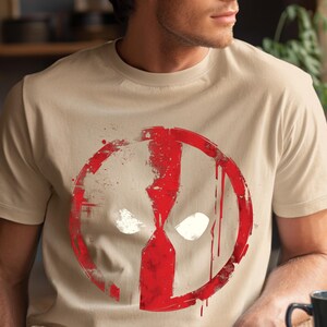 May include: Beige t-shirt featuring a distressed red and white graphic of a character's mask. The design includes a red circle with a white face and two white eye shapes. The shirt is short-sleeved.
