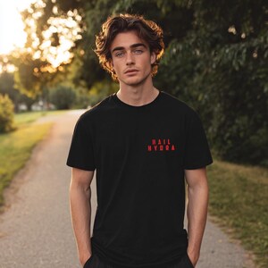 K&ouml;nnte beinhalten: Ein schwarzes kurz&auml;rmeliges T-Shirt mit Rundhalsausschnitt und dem Text "HAIL HYDRA", der in roten Gro&szlig;buchstaben auf der linken Brustseite aufgedruckt ist. Das Kleidungsst&uuml;ck besteht aus einfarbigem schwarzem Stoff und hat eine Standardpassform.