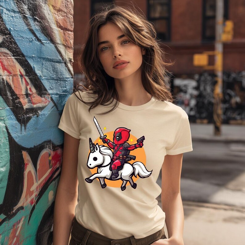 May include: Cream-colored t-shirt featuring a cartoon Deadpool character riding a white unicorn. Deadpool is in a red and black suit, holding a sword and a gun. The design is set against an orange circle.
