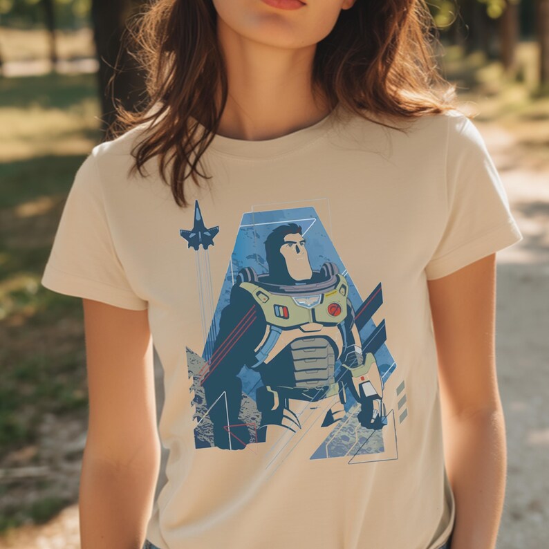 May include: A cream-colored short-sleeve t-shirt featuring a graphic print of a space ranger character in a white, green, and purple suit. The design includes a small blue spaceship, geometric blue shapes, and a textured background element.