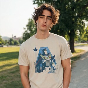 May include: A beige short-sleeved t-shirt featuring a graphic print of a stylized space ranger character in a white, green, and purple suit. The design includes a small rocket ship icon and geometric blue shapes surrounding the central figure.