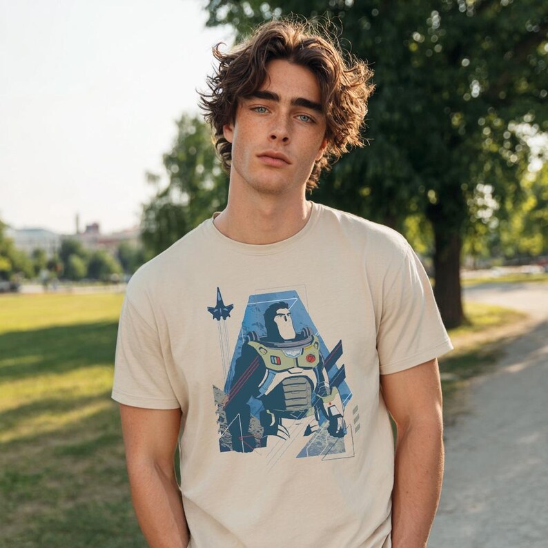 May include: A beige short-sleeved t-shirt featuring a graphic print of a stylized space ranger character in a white, green, and purple suit. The design includes a small rocket ship icon and geometric blue shapes surrounding the central figure.