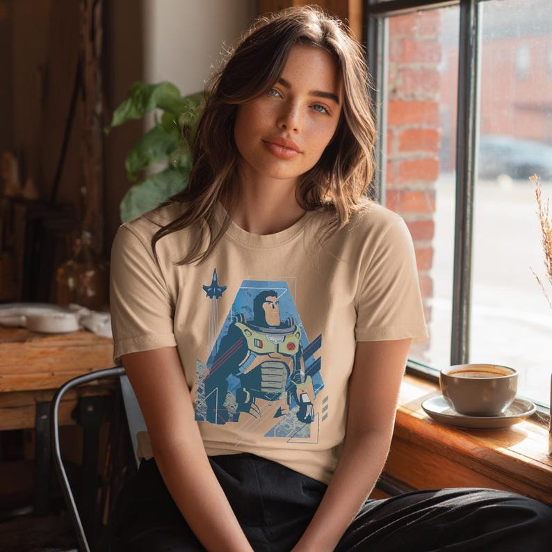 May include: A beige short-sleeved crew neck t-shirt featuring a graphic print of a space ranger character in a white, green, and purple suit, positioned within a blue geometric frame with a small rocket ship icon above the character's head.