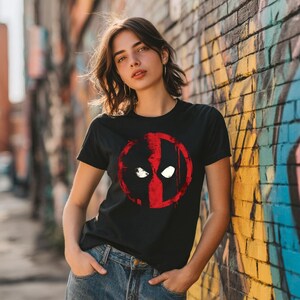 May include: Black t-shirt featuring a red and black graphic of a character's face. The graphic is a stylized design with white eye shapes and a red outline. The t-shirt is a crew neck style.