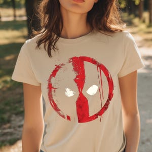 May include: Beige t-shirt featuring a red and white graphic of a character's face. The design is a stylized, circular emblem with a red outline and a central vertical stripe. The eyes are white, and the overall effect is bold and graphic.