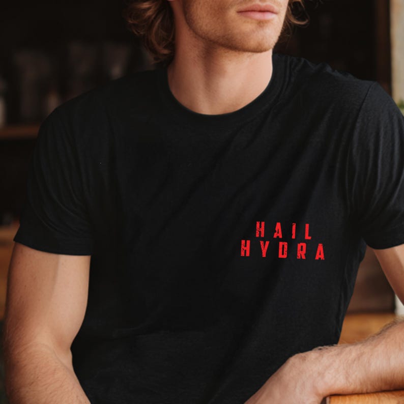 K&ouml;nnte beinhalten: Ein schwarzes T-Shirt mit kurzen &Auml;rmeln und dem Text HAIL HYDRA, der in einer abgenutzten roten Schrift auf der rechten Brustseite aufgedruckt ist. Das Kleidungsst&uuml;ck hat einen klassischen Rundhalsausschnitt und ein schlichtes Design.