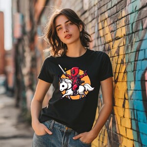 May include: Black t-shirt featuring a cartoon Deadpool character riding a white unicorn. The character is holding a sword and guns, set against an orange circle. The design is a playful mashup of superhero and fantasy themes.