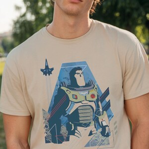 May include: A beige short-sleeved t-shirt featuring a graphic print of a space ranger character in a white, green, and purple suit. The design includes a small blue spaceship flying above the character, set against a blue geometric background.