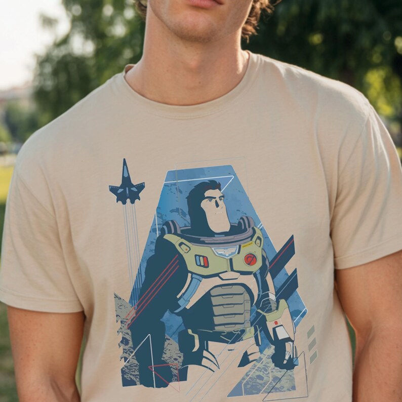 May include: A beige short-sleeved t-shirt featuring a graphic print of a space ranger character in a white, green, and purple suit. The design includes a small blue spaceship flying above the character, set against a blue geometric background.