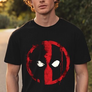 May include: Black t-shirt featuring a red and white graphic of a character's face. The graphic is a stylized design with a circular outline and white eye shapes. The design has a distressed, paint-splatter effect.