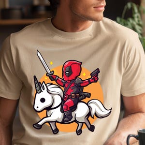 May include: Beige t-shirt featuring a cartoon illustration of Deadpool riding a white unicorn. Deadpool is in his red and black costume, holding a sword and a gun. The unicorn has a white mane and tail, set against an orange circle.