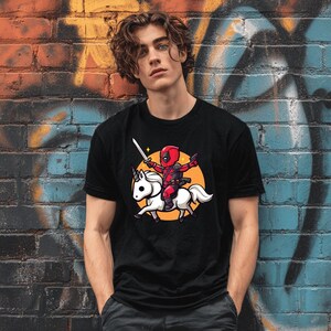 May include: Black t-shirt featuring a cartoon Deadpool character riding a white unicorn. The character is holding a sword and a gun, set against an orange circle. The design is printed on a black t-shirt.