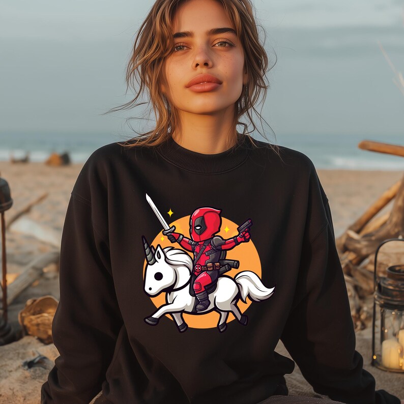 May include: Black sweatshirt featuring a cartoon Deadpool character riding a white unicorn. The character is in a red and black suit, holding a sword and a gun, set against an orange circle. The unicorn has a white mane and tail.