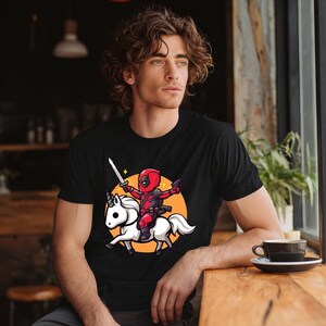 May include: Black t-shirt featuring a cartoon Deadpool character riding a white unicorn. The character is holding a sword and a gun, set against an orange circle. The design is a playful mashup of superhero and fantasy themes.