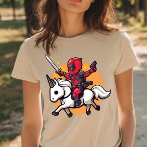 May include: Beige t-shirt featuring a cartoon illustration of a character in a red and black suit riding a white unicorn, holding a sword and a gun. The design includes an orange circle background and star accents.