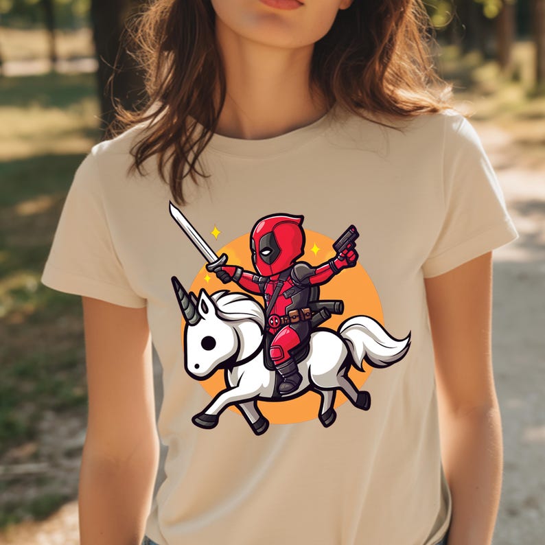 May include: Beige t-shirt featuring a cartoon illustration of a character in a red and black suit riding a white unicorn, holding a sword and a gun. The design includes an orange circle background and star accents.