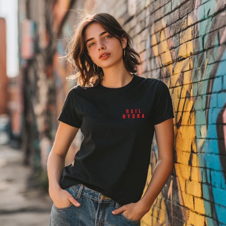 K&ouml;nnte beinhalten: Ein schwarzes Kurzarm-T-Shirt mit dem roten Schriftzug "HAIL HYDRA" auf der linken Brustseite. Das T-Shirt hat einen Rundhalsausschnitt und wird mit einer blauen Jeanshose kombiniert, die sichtbare Metallkn&ouml;pfe und Taschen aufweist.