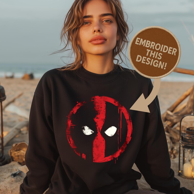 May include: Black sweatshirt featuring a red and white graphic design. The design is a stylized face with white eyes and a red outline. The text "EMBROIDER THIS DESIGN!" is on a brown patch.