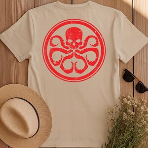 K&ouml;nnte beinhalten: Ein beiges Kurzarm-T-Shirt mit einer gro&szlig;en, kreisf&ouml;rmigen roten Grafik auf der R&uuml;ckseite. Das Design zeigt einen verwitterten Totenkopf in einem Kreis, der von mehreren tentakelartigen Armen umgeben ist, die nach au&szlig;en ragen.