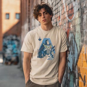 May include: A beige short-sleeved t-shirt featuring a graphic print of a space ranger character in a white, green, and purple suit. The design includes a small rocket ship icon and geometric blue shapes, centered on the chest of the garment.