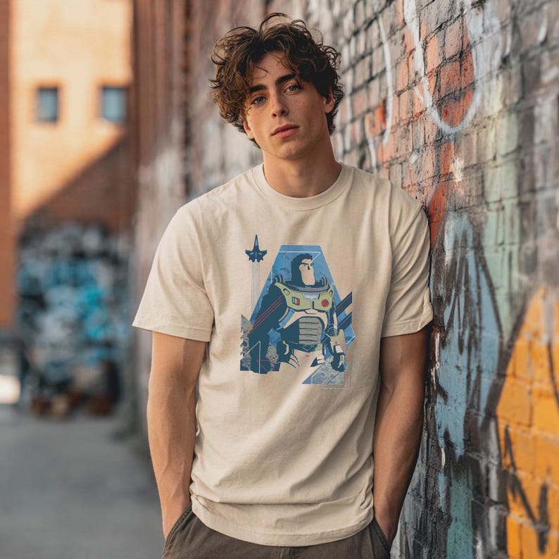 May include: A beige short-sleeved t-shirt featuring a graphic print of a space ranger character in a white, green, and purple suit. The design includes a small rocket ship icon and geometric blue shapes, centered on the chest of the garment.