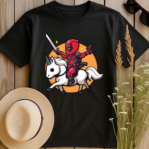 May include: Black t-shirt featuring a cartoon Deadpool character riding a white unicorn. Deadpool is in his red and black suit, holding a sword and a gun. The design is set against an orange circle.