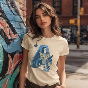 May include: A cream-colored short-sleeve t-shirt featuring a graphic print of a space ranger character in a blue and white suit, with a small rocket ship icon above the character, all set within a geometric blue frame on the front of the shirt.