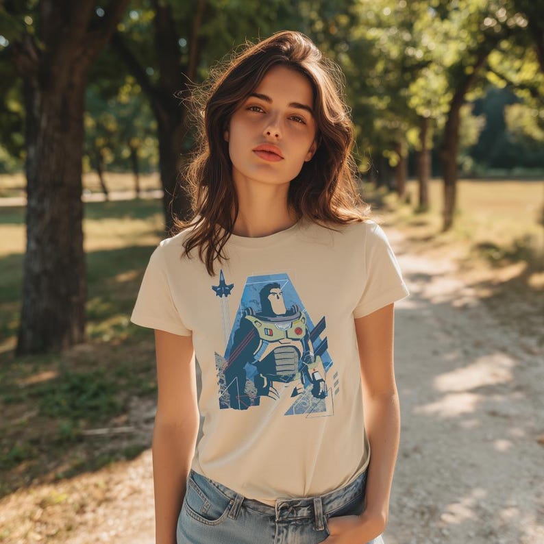 May include: A cream-colored short-sleeve t-shirt featuring a graphic print of a space ranger character in a white, green, and purple suit. The design includes blue geometric shapes and a small rocket ship icon positioned above the character's head.