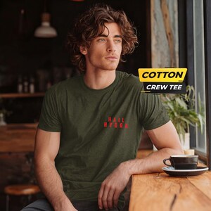 K&ouml;nnte beinhalten: Ein olivgr&uuml;nes Kurzarm-T-Shirt mit Rundhalsausschnitt und dem roten Schriftzug "HAIL HYDRA" auf der linken Brustseite. Eine gelb-schwarze Grafik in der oberen rechten Ecke zeigt den Text "COTTON CREW TEE".