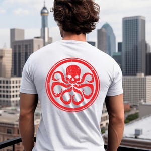K&ouml;nnte beinhalten: Ein wei&szlig;es Kurzarm-T-Shirt mit einer gro&szlig;en, kreisf&ouml;rmigen roten Grafik auf der R&uuml;ckseite. Das Design zeigt einen stilisierten Totenkopf in einem Ring, umgeben von mehreren tentakelartigen Formen, alles in einer abgenutzten, verwitterten Optik.